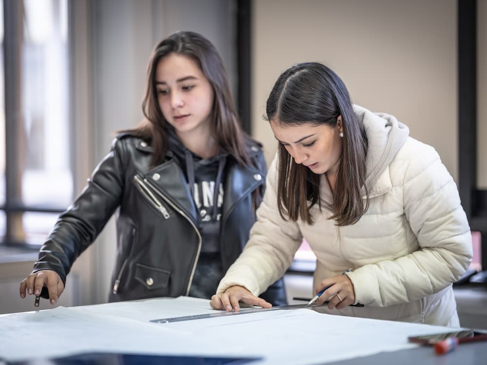 Dos estudiantes trabajando juntas sobre plano o patrón técnico en aula, usando compás y reglas.