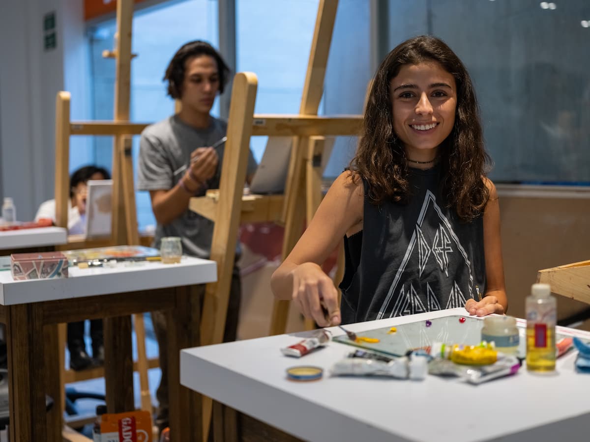 Estudiante sonriendo mientras prepara su paleta de colores en clase de pintura con caballetes al fondo.