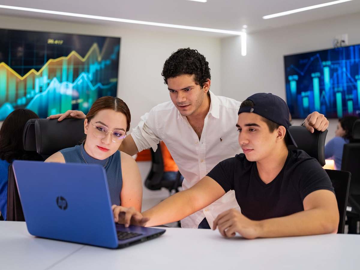 Grupo de estudiantes analizando datos en una laptop en aula tecnológica, con pantallas que muestran gráficos bursátiles en tiempo real.