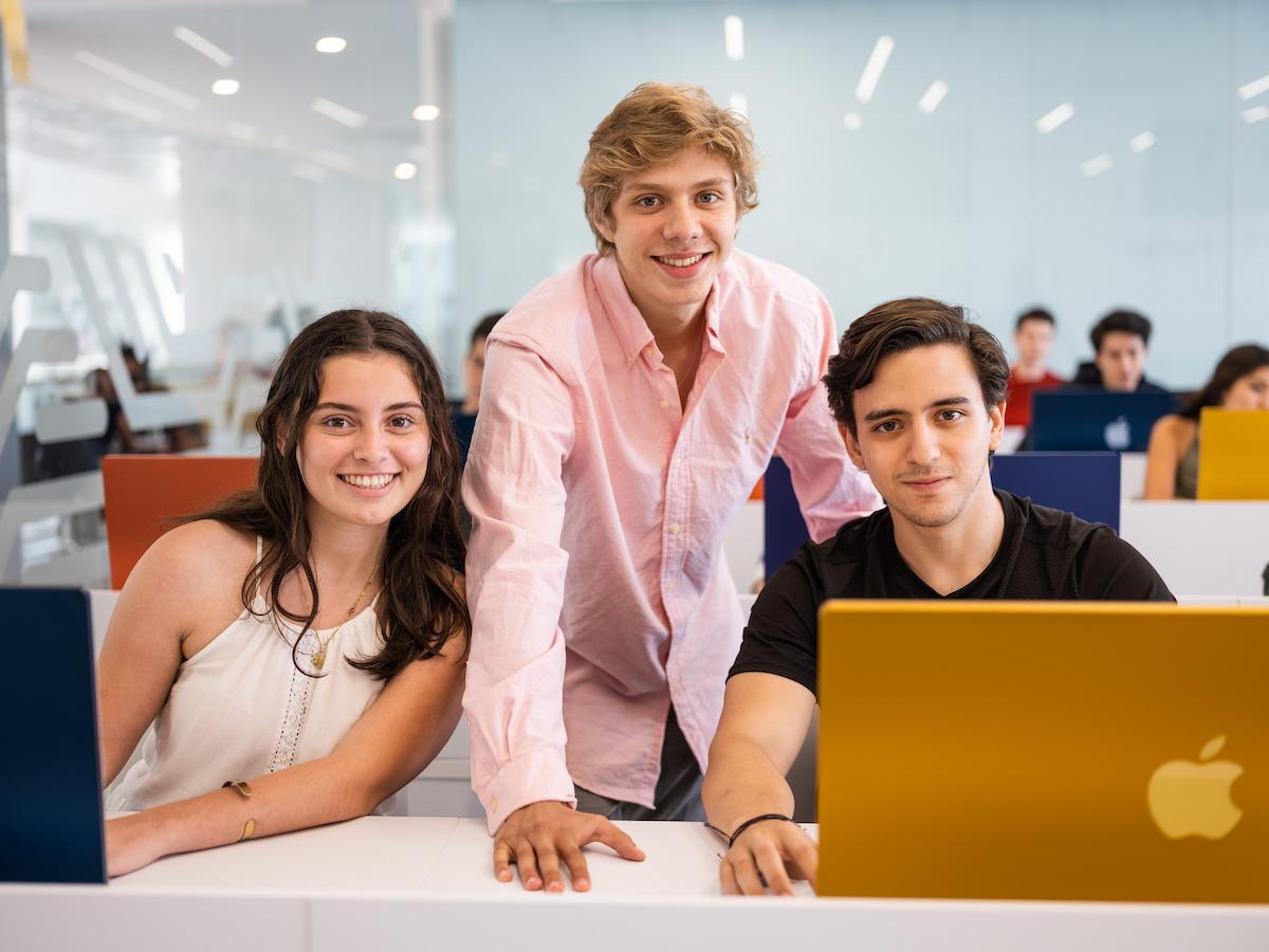 Dos estudiantes sentados y uno de pie en un aula con laptops de colores, sonriendo y proyectando entusiasmo por el aprendizaje en la Universidad Anáhuac.