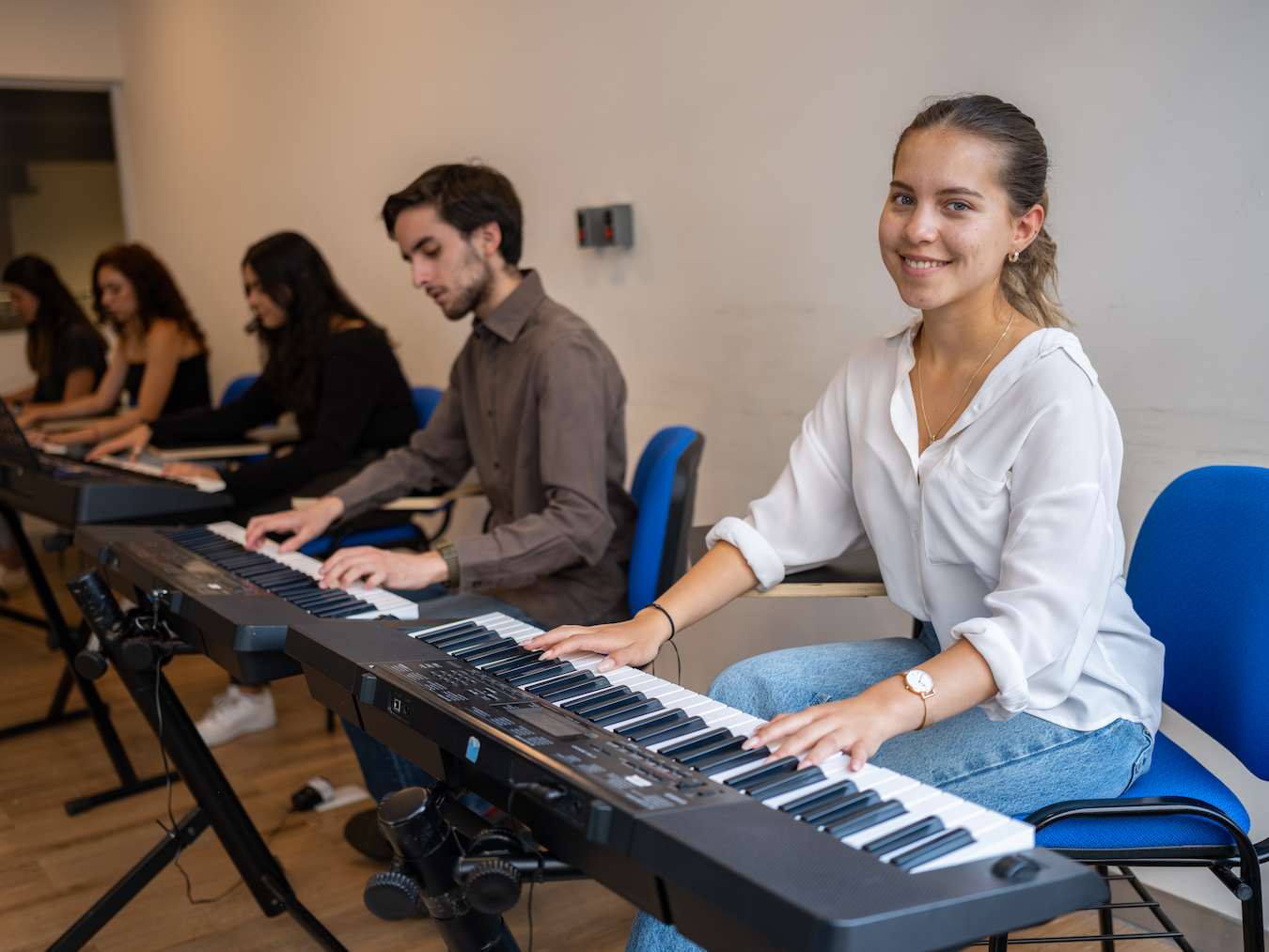 Estudiantes sentados en fila tocando teclados electrónicos durante una clase de piano, en un aula con piso de madera; al frente, una joven con camisa blanca y jeans sonríe a la cámara mientras toca su instrumento.