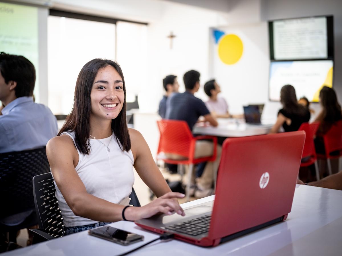 Estudiante de Pedagogía Organizacional y Educativa usando su laptop y sonriendo en un aula dinámica, reflejando aprendizaje activo, uso de tecnología y formación en entornos educativos y organizacionales.