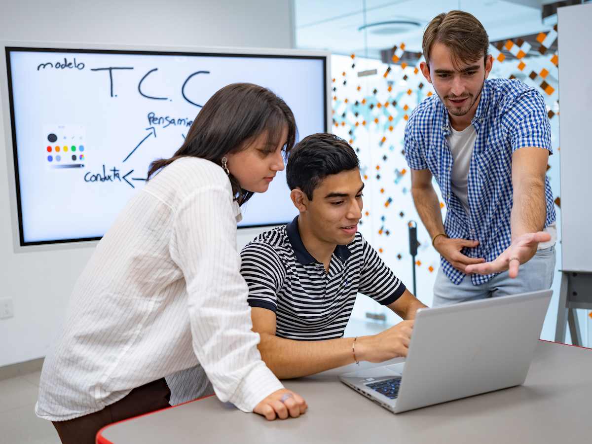 Estudiantes de Psicopedagogía colaborando frente a una laptop con una pizarra digital al fondo que muestra el modelo TCC, reflejando análisis cognitivo, trabajo en equipo y aplicación de teorías del aprendizaje.