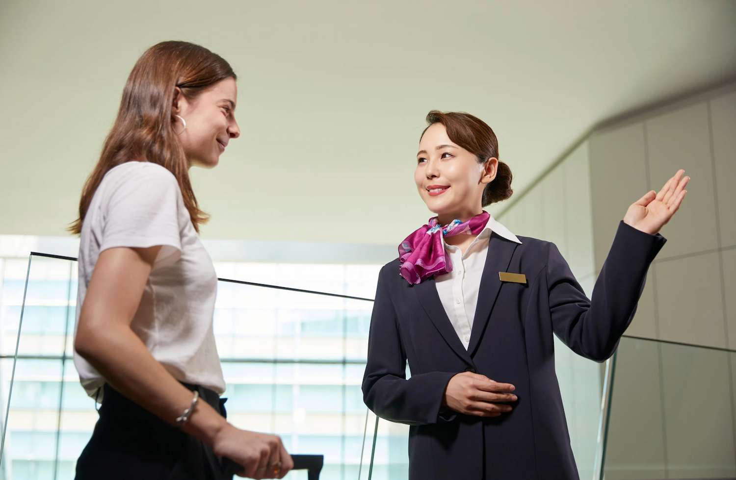 Empleada de hotel con uniforme formal y mascada fucsia sonríe mientras indica una dirección a una visitante, en un ambiente moderno e iluminado.