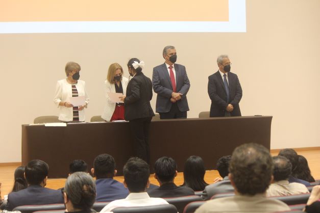 Un presidium con dos mujeres y dos hombres trajeados, entregando un reconocimiento a una adolescente.