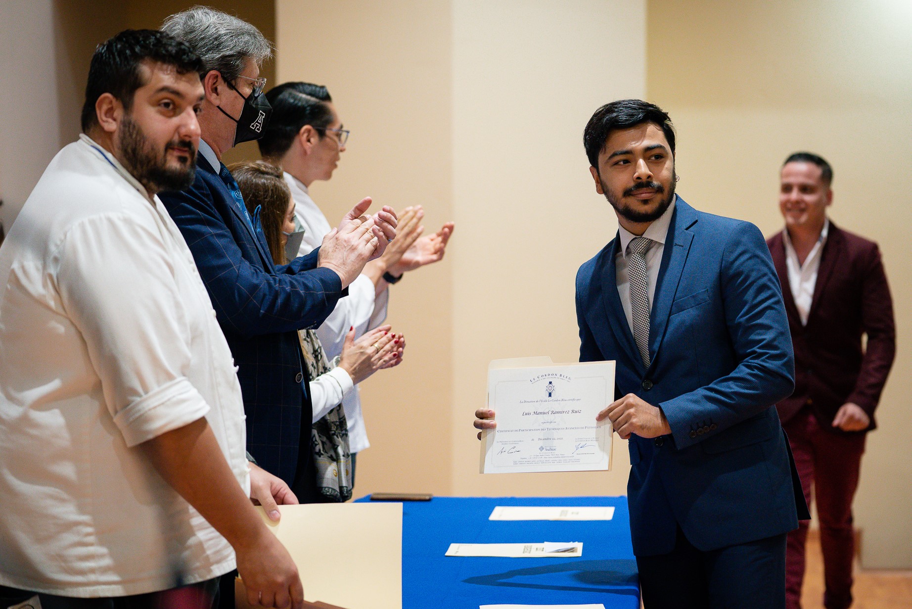 Alumno moreno, de barba y con traje azul recibe reconocimiento de Le Cordon Bleu.