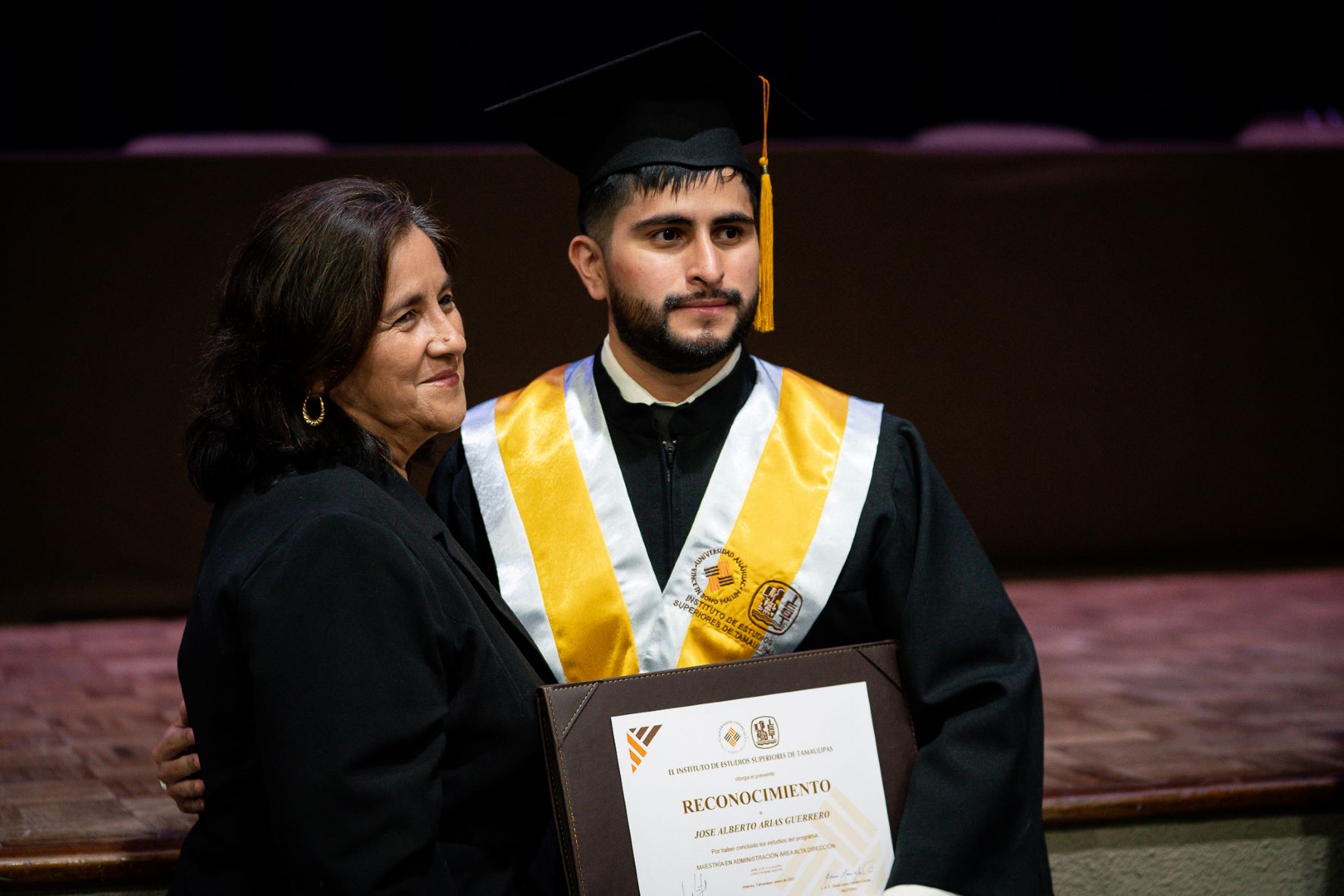 Joven de barba con toga y birrete, en compañía de su mamá, mostrando su título.