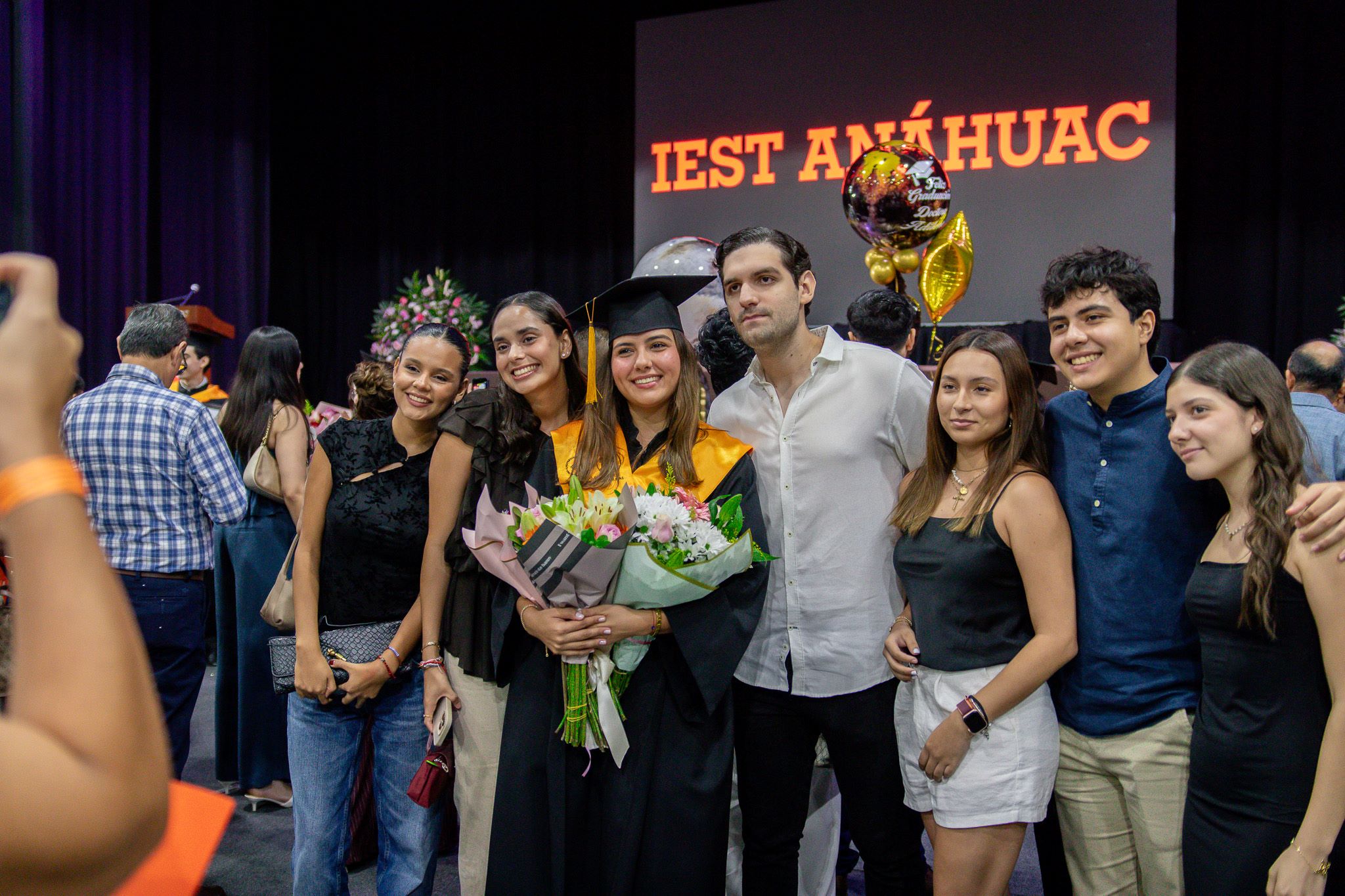 Una familia posa con su graduada, mientras les toman una foto en el Gimnasio Burton Grossman. Atrás dice la frase: IEST Anáhuac.