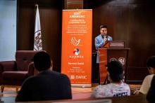 Un alumno de cabello corto habla a una audiencia. Atrás tiene un banner naranja.