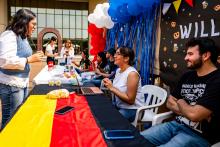 Una alumna habla con otra, sobre una mesa con la bandera de Alemania, mientras un chico de barba la observa.