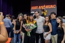 Una familia posa con su graduada, mientras les toman una foto en el Gimnasio Burton Grossman. Atrás dice la frase: IEST Anáhuac.